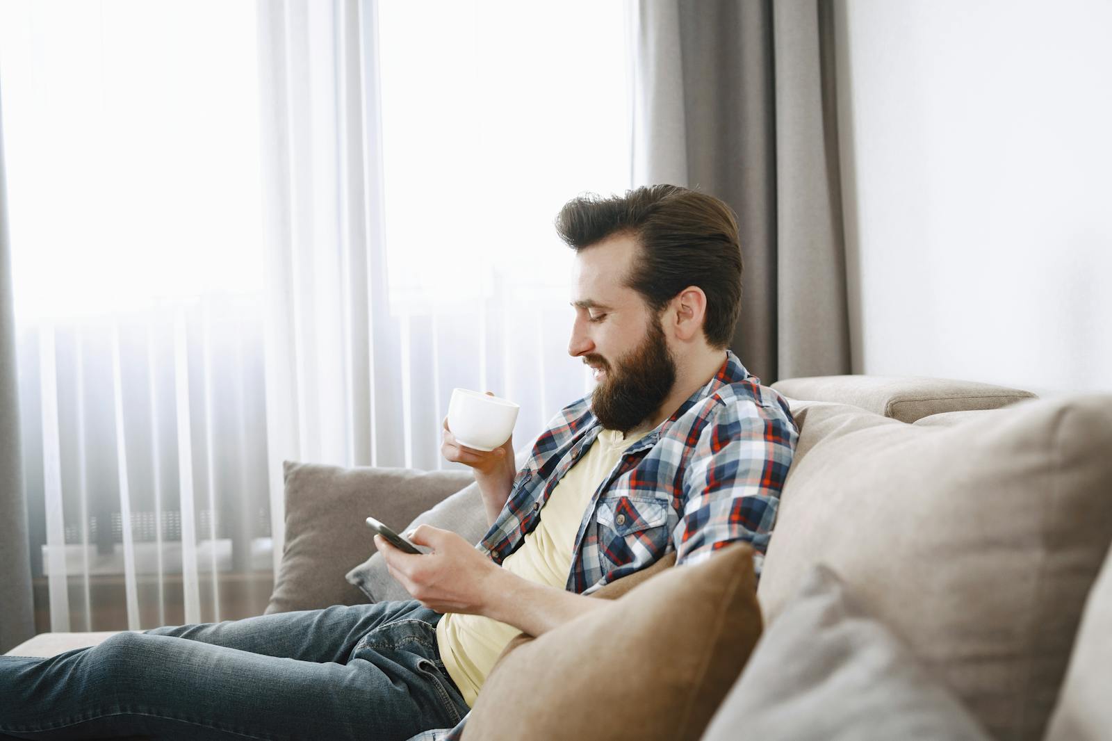 Casual bearded man enjoying coffee on a comfy sofa while browsing on his phone.